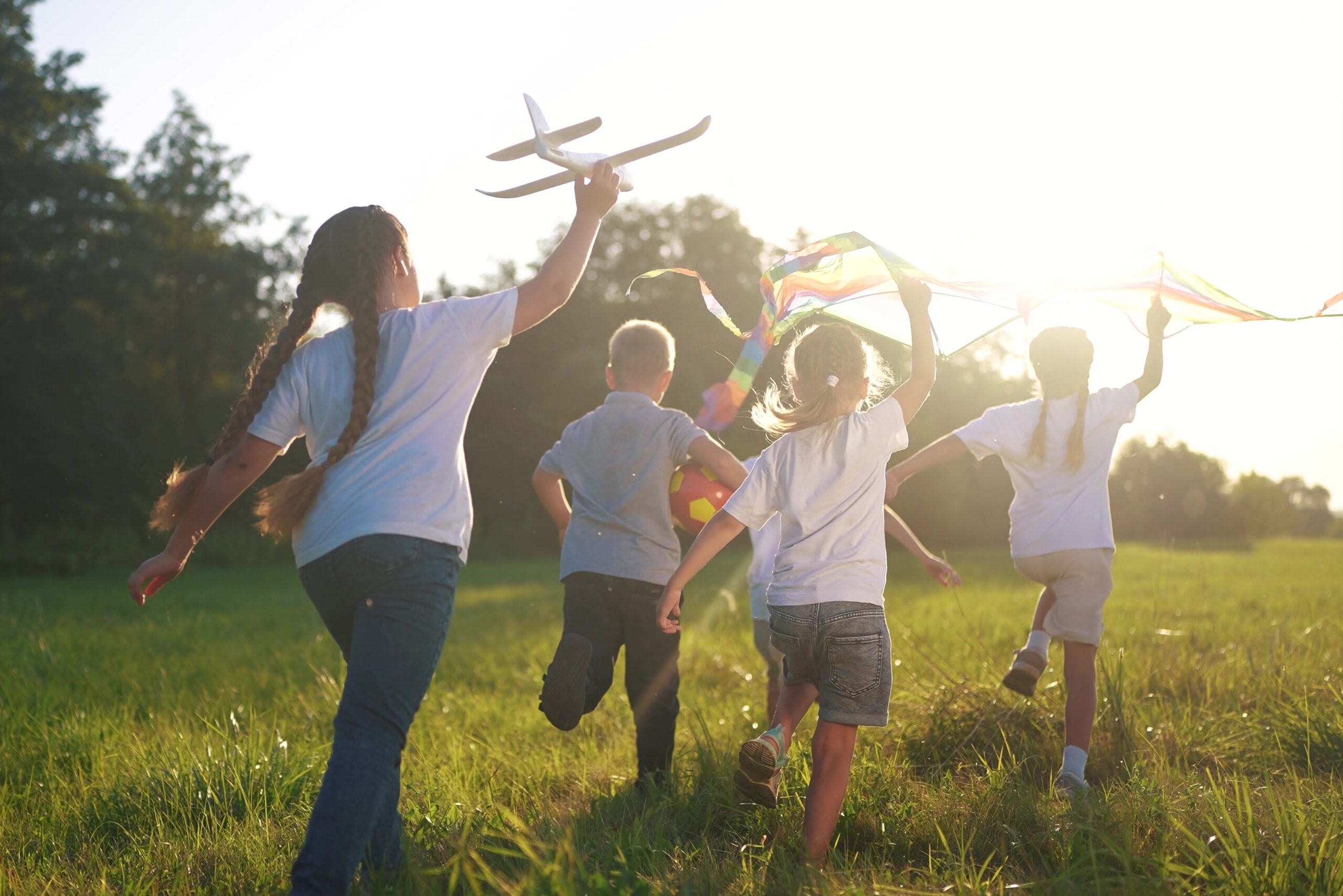 Das Bild zeigt Kinder von hinten, die auf einer Wiese rennen mit einem Modellflugzeug und Luftschlangen.