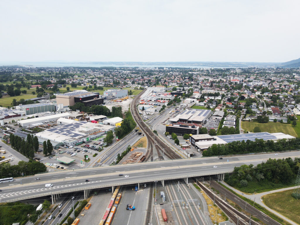 Aufnahme einer Drohne über das Industriegebiet von Lauterach. Man sieht eine Autobahnbrücke, viele Firmengebäude und Firmengelände und eine Straße, die durch die Gemeinde führt.