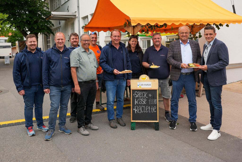 Das Bild zeigt eine Gruppe vor dem Stand der Feuerwehr mit Speck- und Käsetellern. Alle lachen in die Kamera, im Hintergrund ein gelbes Zeltdach.

