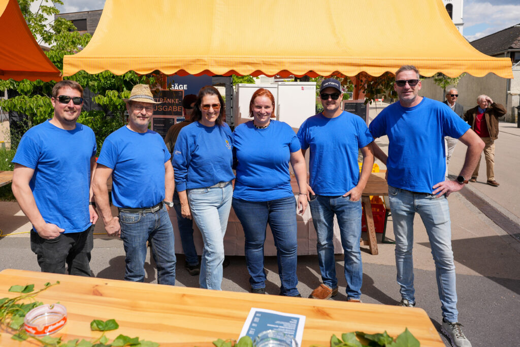 Das Bild zeigt sechs Personen der Gilde Unterfeld in blauen T-Shirts vor einem Getränkestand. Sie stehen unter einem gelben Zelt und posieren gut gelaunt für die Kamera.