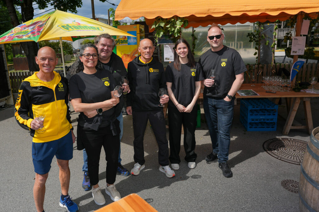 Das Bild zeigt ein Team des Karate Hofsteig, das mit leeren Weingläsern unter einem Stand steht. Alle tragen schwarze Kleidung mit gelbem Vereinslogo.