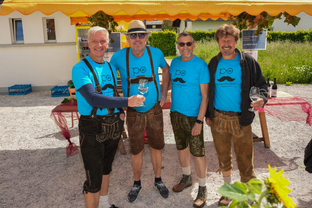 Das Bild zeigt fünf Männer des Männerchors Lauterach in blauen T-Shirts und Lederhosen. Sie halten Weingläser und stehen vor einem Verkaufsstand.