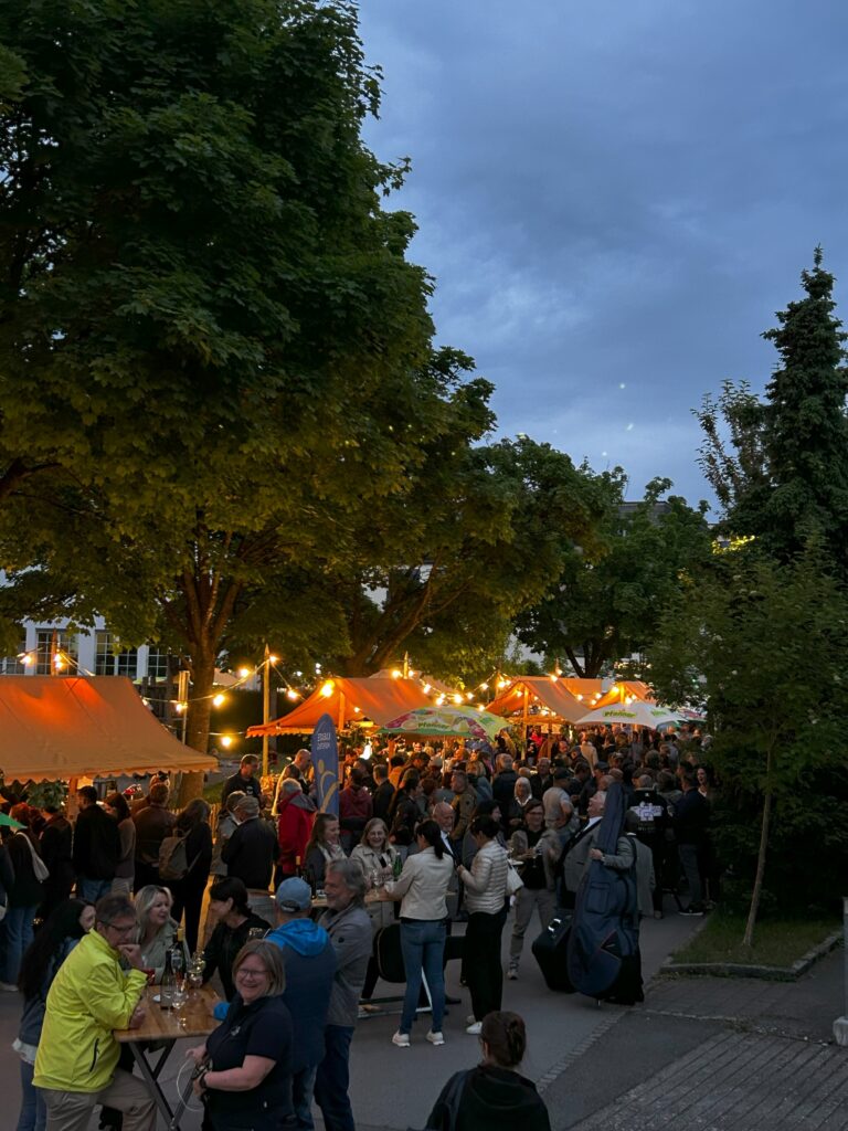 Das Bild zeigt das abendliche Festgelände bei der Säge Lauterach. Viele Menschen stehen bei den beleuchteten Ständen unter Bäumen und genießen die Stimmung.