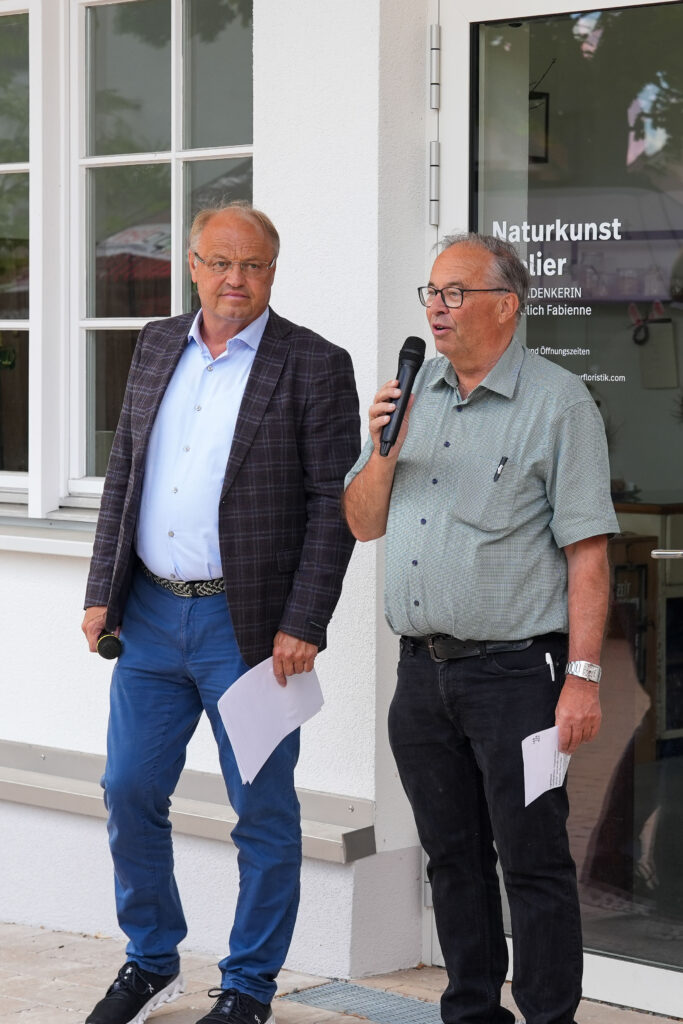 Das Bild zeigt den Bürgermeister Elmar Rhomberg und Walter Pfanner bei der offiziellen Eröffnung vor dem Naturkunst-Atelier. Einer spricht ins Mikrofon, beide halten Blätter mit Notizen.