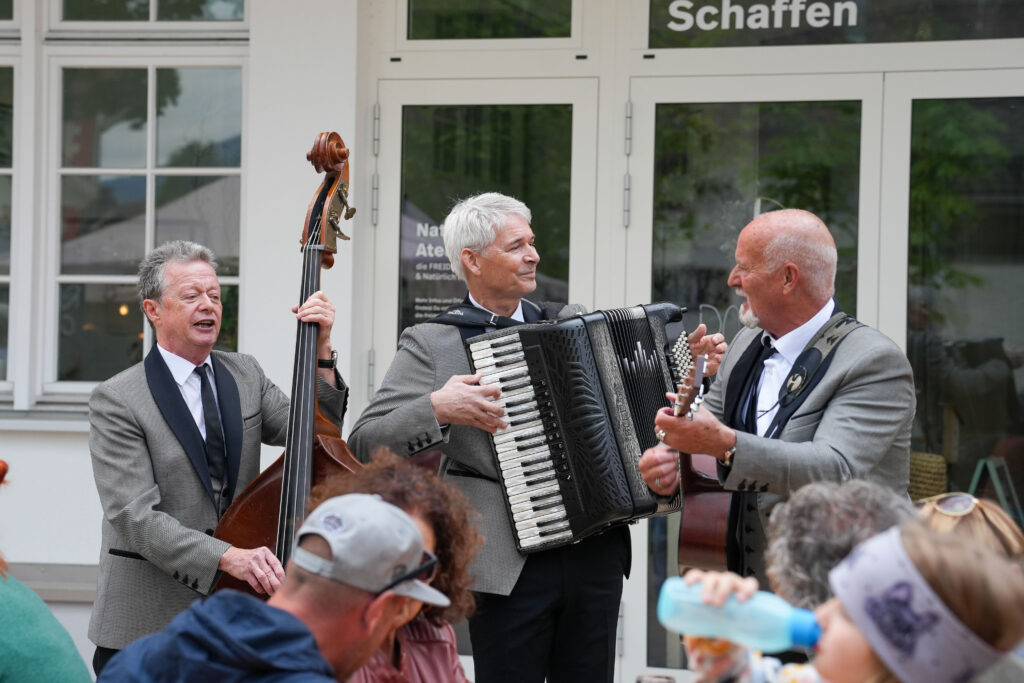 Das Bild zeigt drei Musiker beim Auftritt vor dem Naturkunst Atelier. Sie spielen Kontrabass, Akkordeon und Gitarre und sorgen für musikalische Begleitung beim Fest.