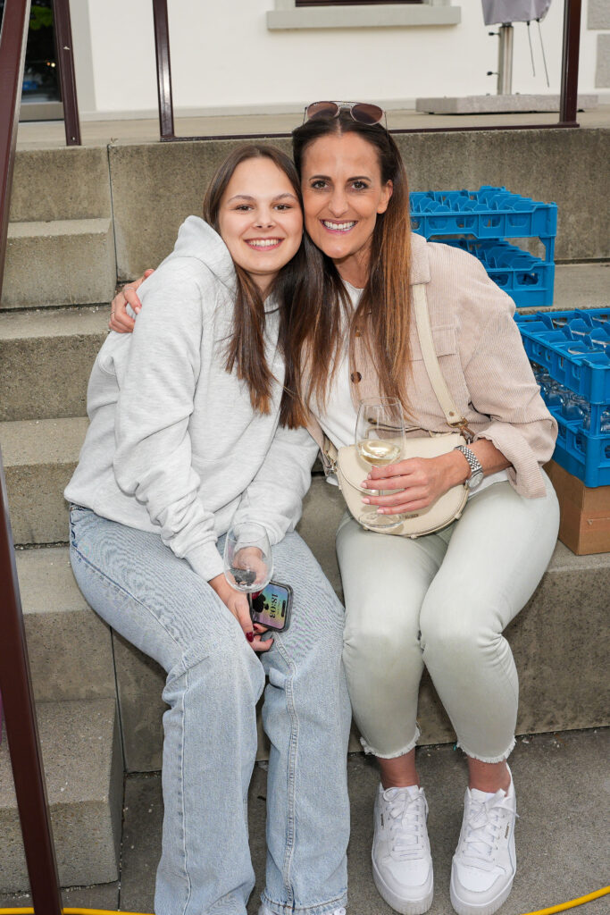 Das Bild zeigt zwei Frauen, die auf einer Stufe sitzen und sich umarmen. Beide lachen in die Kamera und halten Weingläser in der Hand.