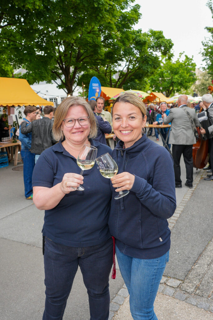 Das Bild zeigt das Veranstaltungsteam beim Anstoßen mit Weißwein. Im Hintergrund ist das bunte Festgelände mit vielen Gästen und Ständen zu sehen.