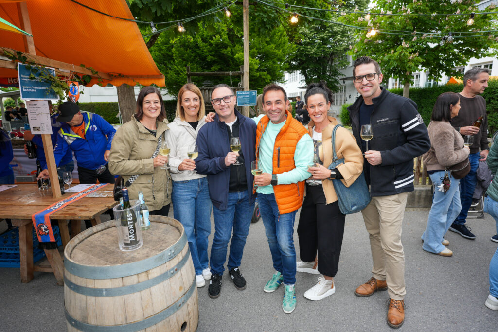 Das Bild zeigt eine sechsköpfige Gruppe, die mit Weingläsern vor einem Stand posiert. Alle lachen in die Kamera, das Festgelände ist gut besucht.