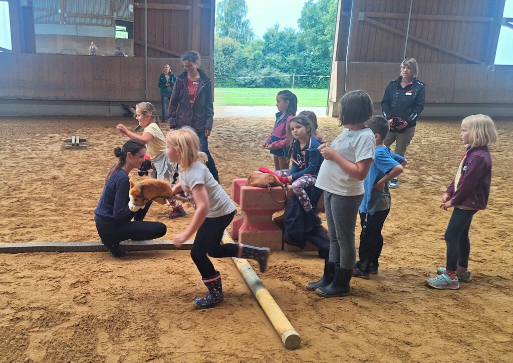 Das Bild zeigt Kinder in einer Reithalle. Ein Kind reitet auf einem Holzpferd.