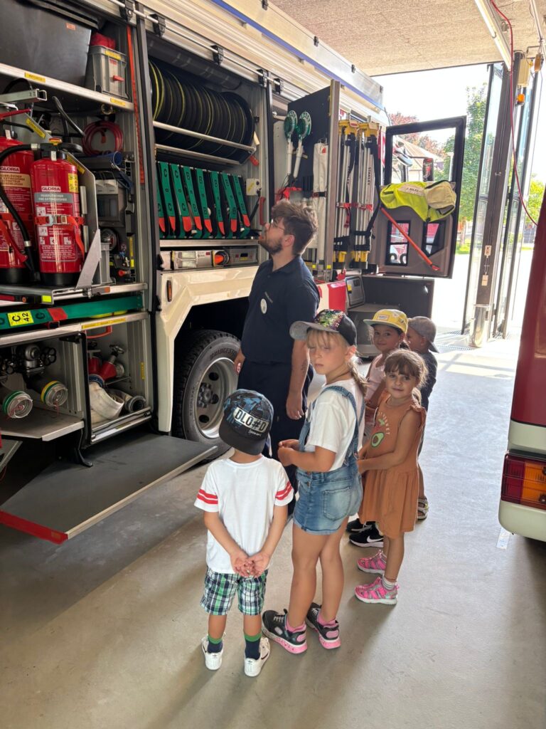 Das Bild zeigt einen Feuerwehrmann, der vier Kindern die Geräte in einem Feuerwehrauto erklärt.