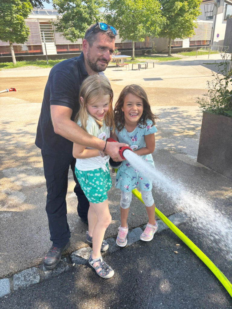 Das Bild zeigt einen Feuerwehrmann, der zwei Kindern beim Zielspritzen mit dem Feuerwehrschlauch hilft.