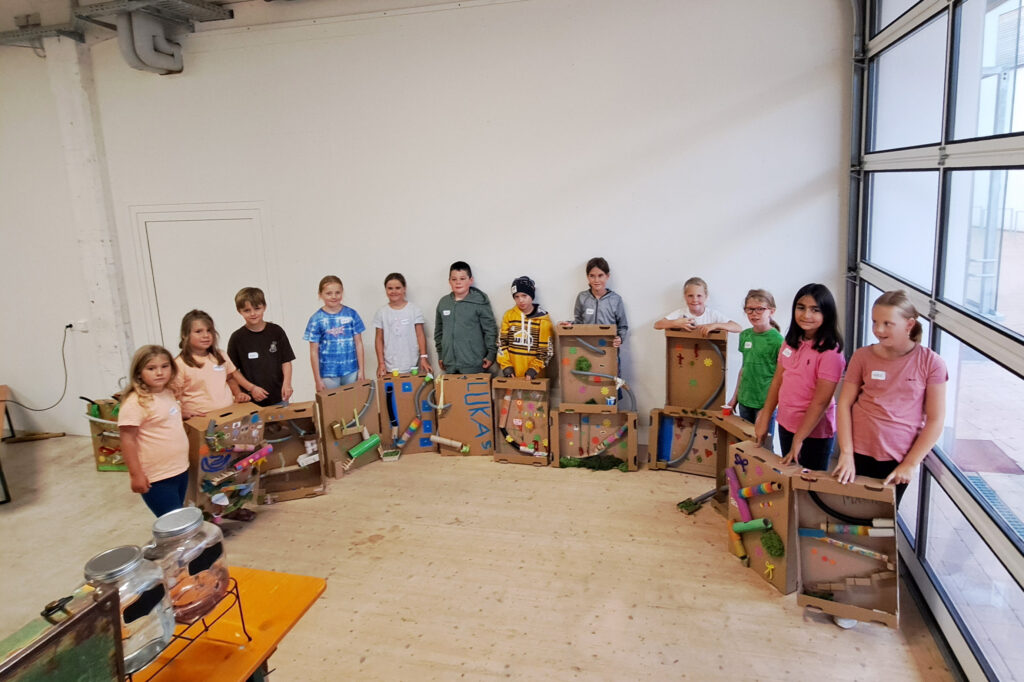 Das Bild zeigt eine Gruppe von Kindern, die ihre selbstgebastelten Kugelbahnen aus Klopapierrollen präsentieren.