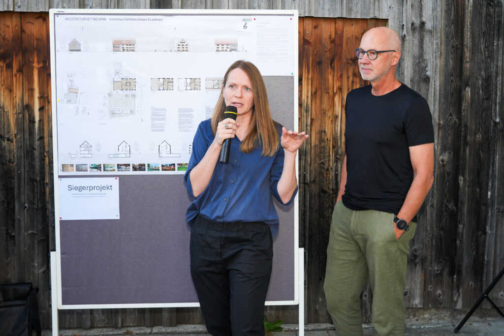 Die Architekten Claudia Greußing und Christoph Kalb stehen vor dem Plänen des neuen Kinderhauses und sprechen in ein Mikrofon.