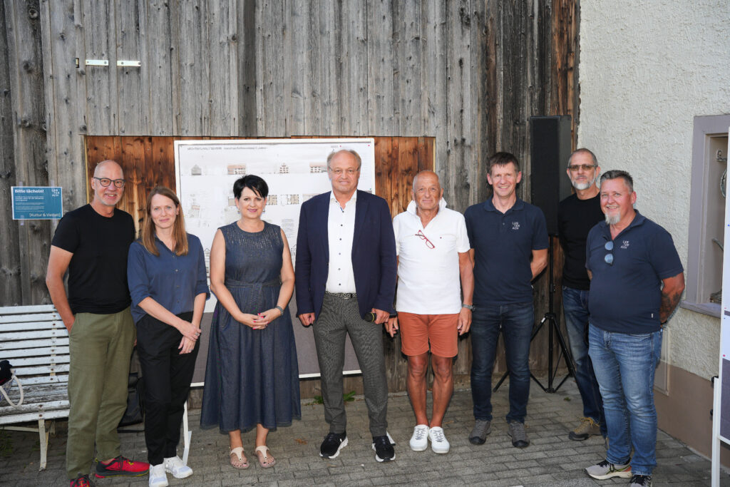 Eine Gruppe von Menschen steht vor den Plänen des Fesslerhauses: Architekt Christoph Kalb und Architektin Claudia Greußing, Vizebürgermeisterin und Vorsitzende des Bildungsauschusses Sabine Kassegger, Bürgermeister Elmar Rhomberg, ehemaliger Eigentümer Felix Fessler, Baumeister Herbert Willam (Fachbereichsleiter Baurecht und Hochbau der MG Lauterach), Architekt Andreas Flatz und Jürgen Ropele (Bauleiter der MG Lauterach)