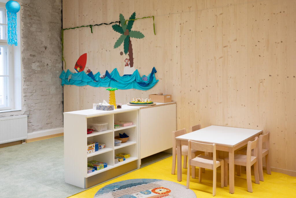 Das Bild zeigt einen Kindergartenraum mit kleiner Sitzecke, weißen Regalen voller Spiele und bunter Wanddeko mit Palme, Meer und Boot.