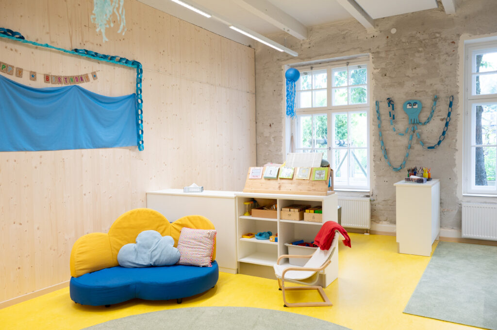 Das Bild zeigt einen hellen Kindergartenraum mit gemütlicher Sofa-Ecke, Regalen voller Spielsachen und einer Wanddeko mit blauem Oktopus und Geburtstagsgruß.