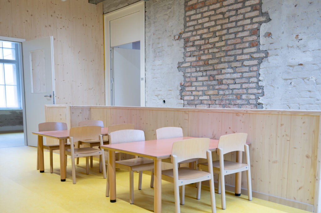 Das Bild zeigt einen Kindergartenraum mit kleinen Holztischen und Stühlen, gelbem Boden und einer Wand mit sichtbarem Backstein und Holzverkleidung.