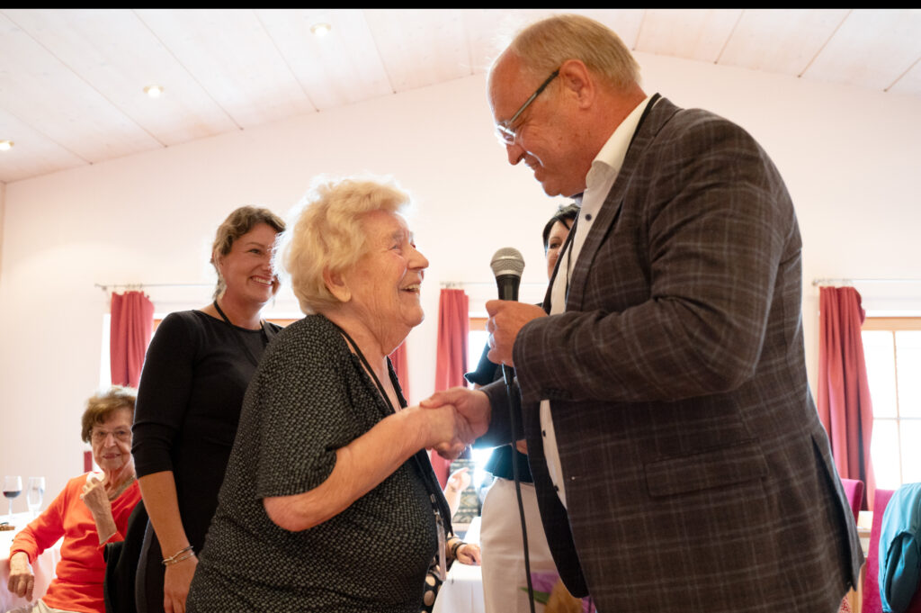 Das Bild zeigt eine Seniorin, die bei einer Feier einem Mann die Hand schüttelt, er hält ein Mikrofon.