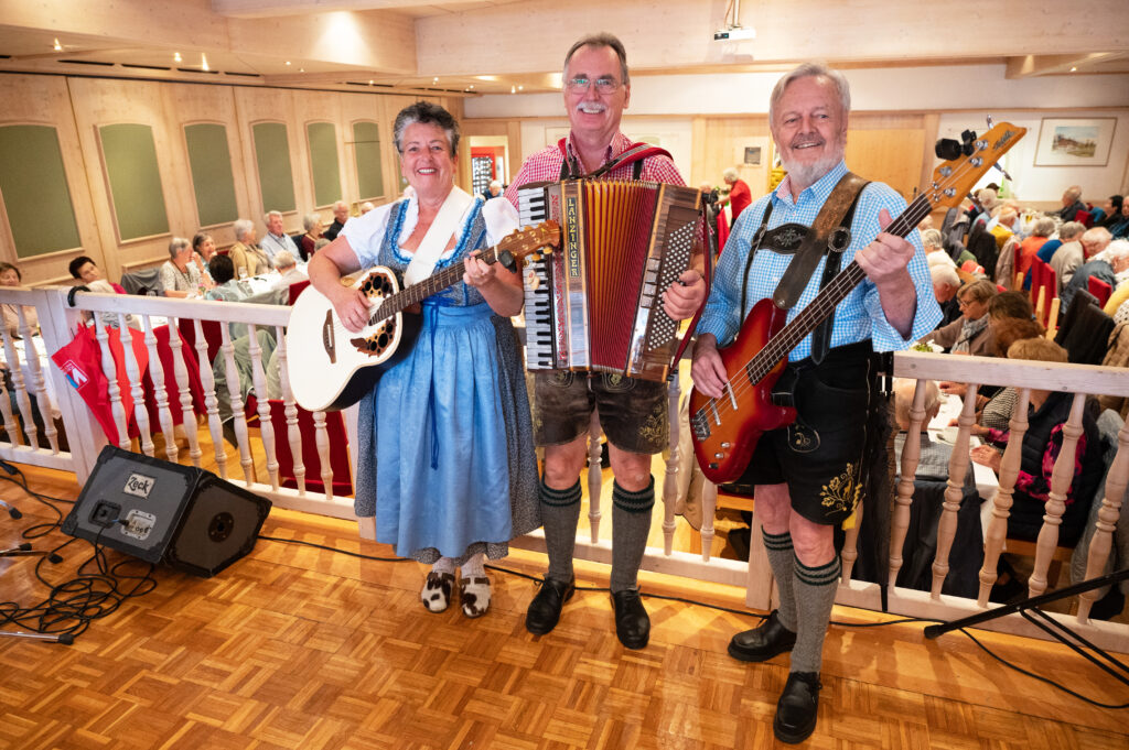 Das Bild zeigt drei Musiker:innen in Tracht mit Gitarre, Akkordeon und Bass vor einem Publikum.