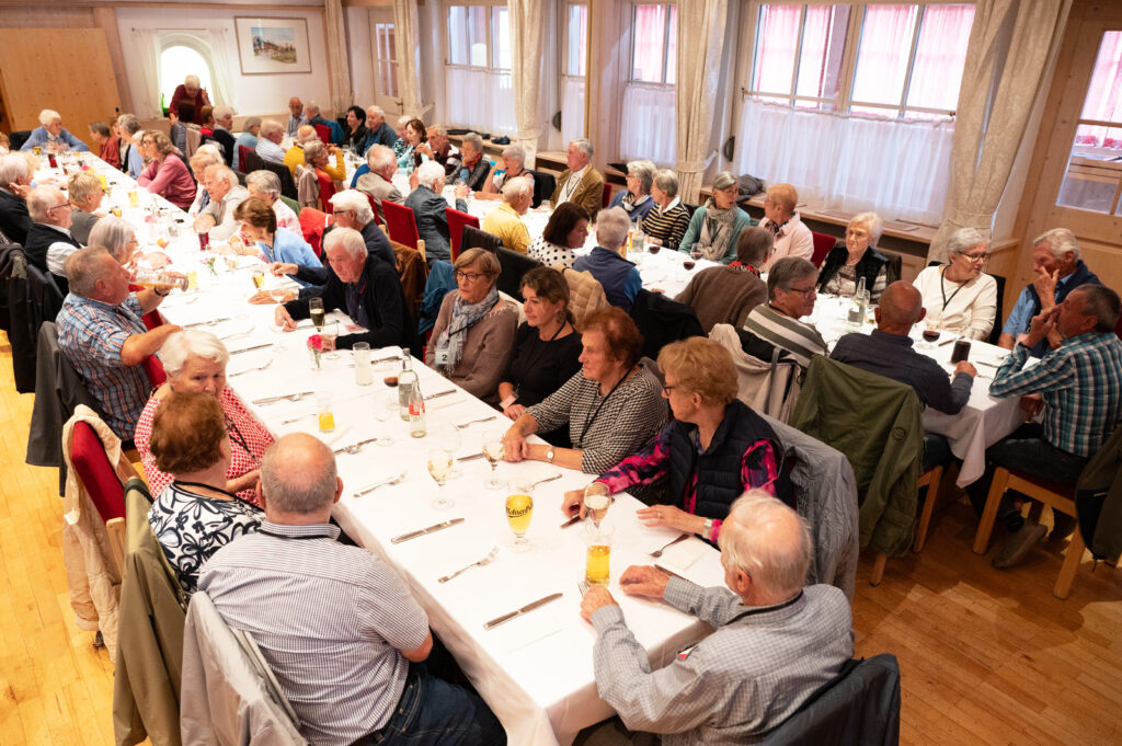 Das Bild zeigt viele Senior:innen, die an langen Tischen in einem hellen Festsaal zusammensitzen.