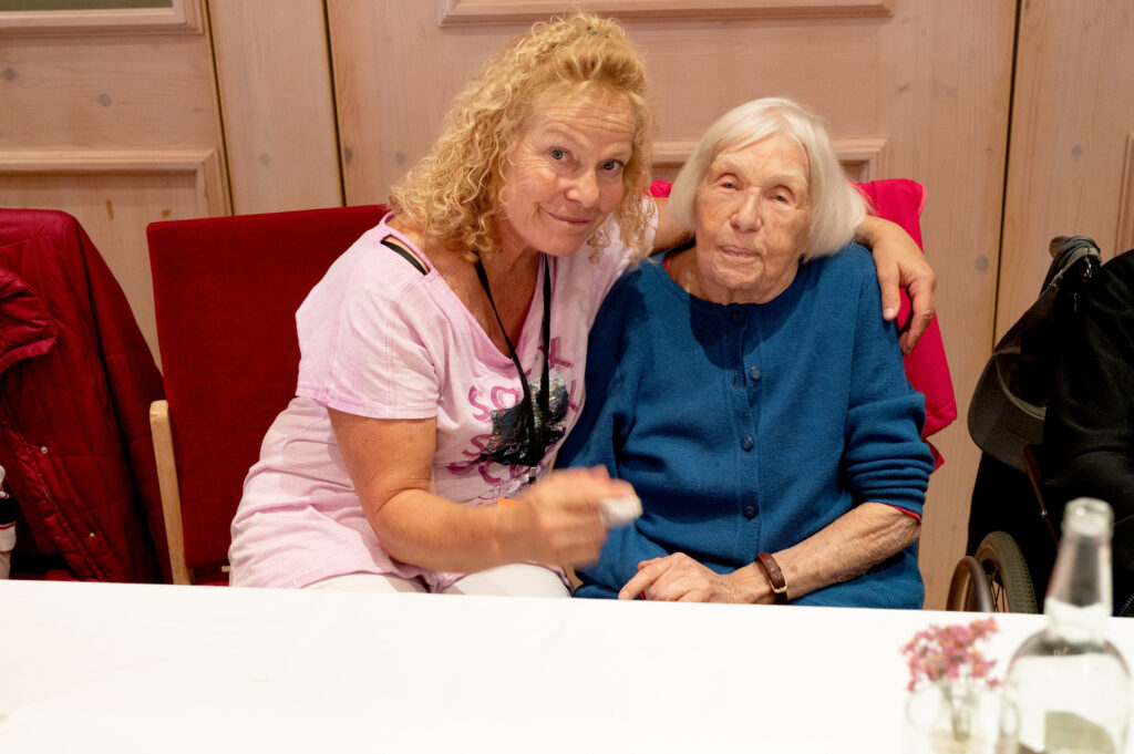 Das Bild zeigt zwei Frauen, eine davon im Rollstuhl, die sich bei einer Feier umarmen.