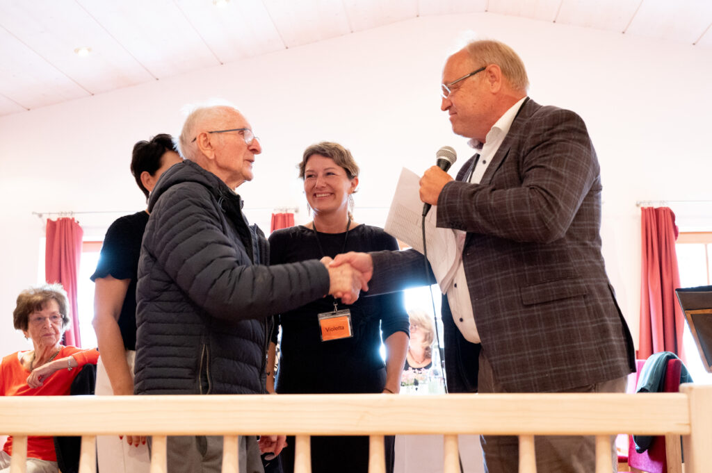 Das Bild zeigt Josef Berkmann, der Bürgermeister Elmar Rhomberg mit Mikrofon die Hand schüttelt, zwei Frauen stehen daneben.