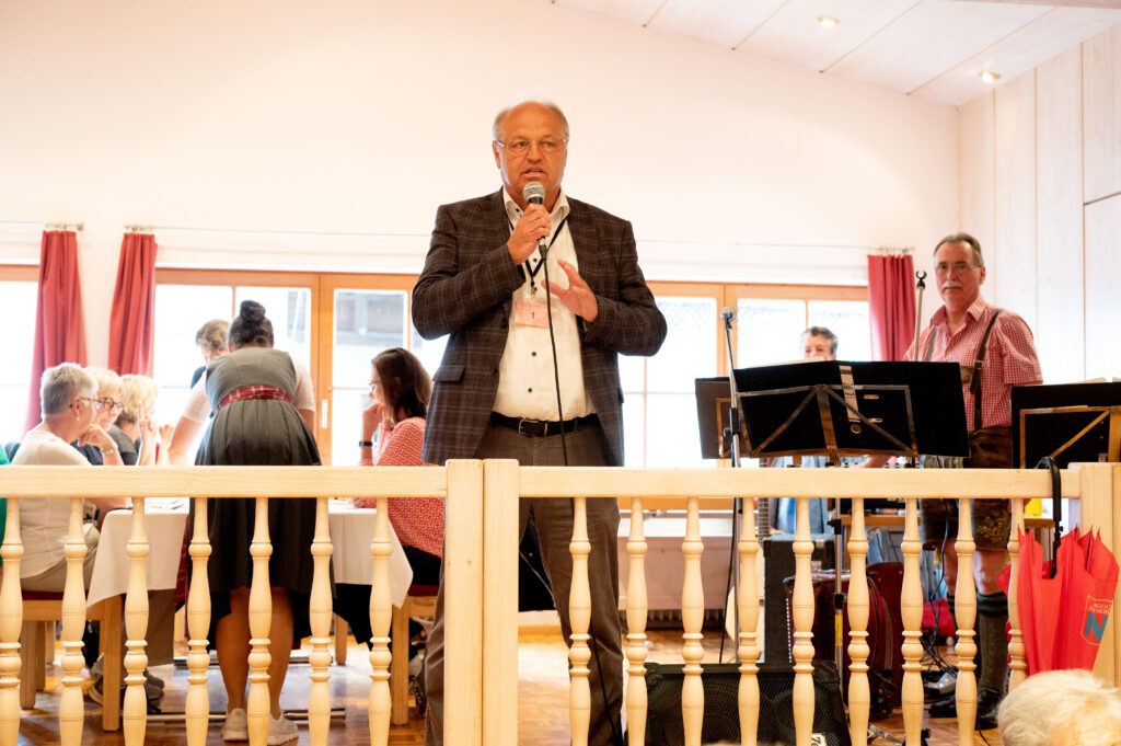 Das Bild zeigt Bürgermeister Elmar Rhomberg mit Mikrofon, der zu den Gästen spricht, im Hintergrund Musikanten.
