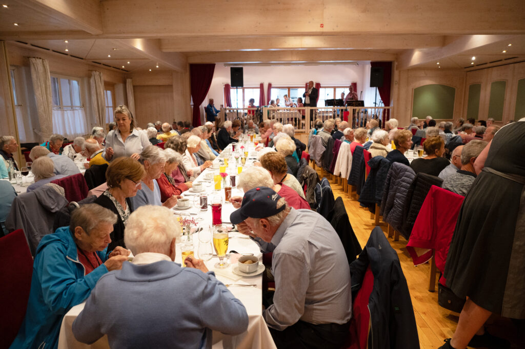 Das Bild zeigt viele Senior:innen an langen Tischen bei einem Festessen im großen Saal.