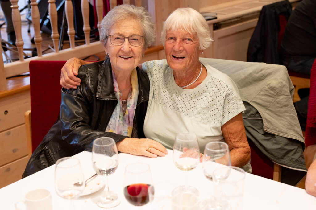 Das Bild zeigt zwei Seniorinnen, die sich bei einem Festessen umarmen und in die Kamera lächeln.