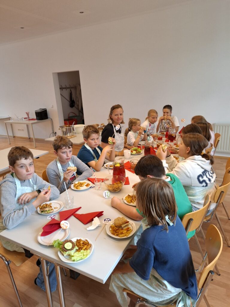 Das Bild zeigt eine Gruppe von Kindern, die ihr selbstgemachtes Fastfood am Tisch essen.
