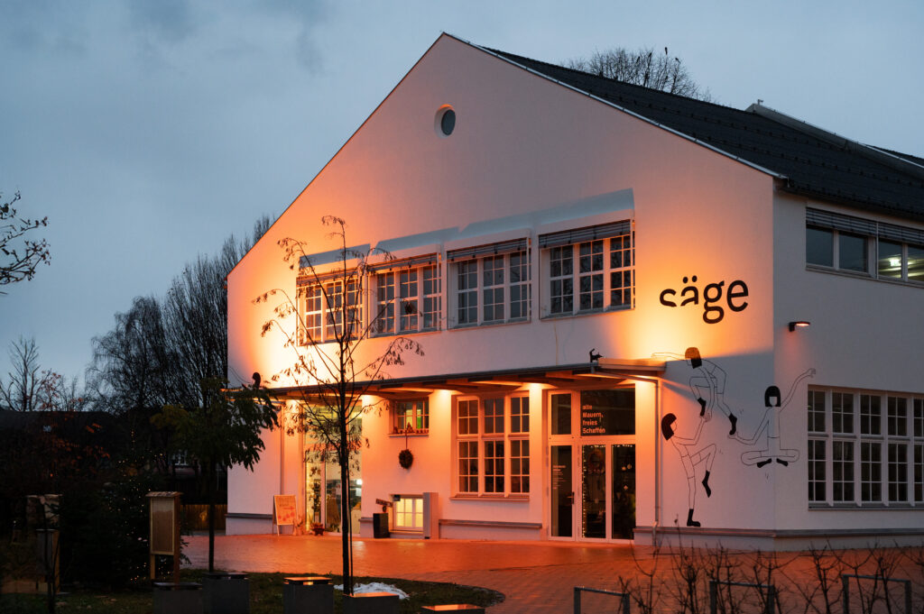 Das Bild zeigt die Säge in Lauterach am Abend, orange beleuchtet zu den Aktionstagen "Orange the World – 16 Tage gegen Gewalt an Frauen".