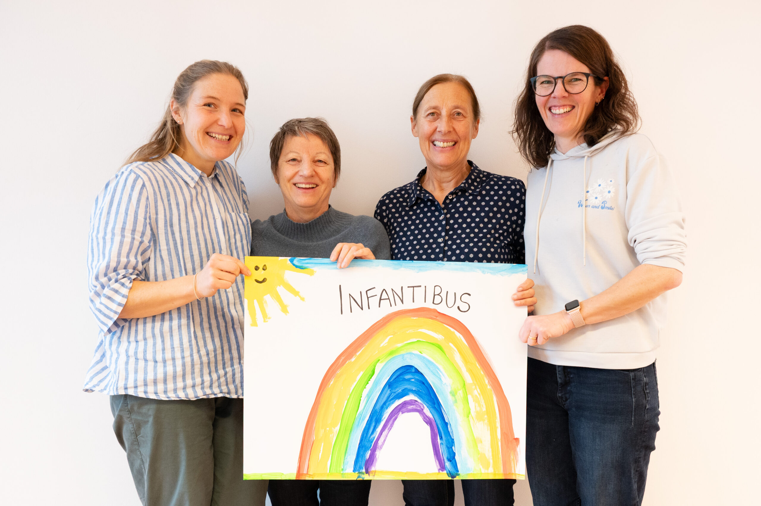 Das Bild zeigt ehemalige und aktuelle Frauen des Infantibus-Teams, die ein Plakat mit einem Regenbogen in die Kamera halten.