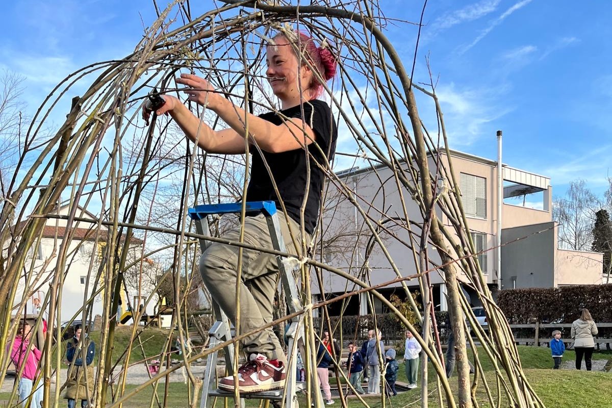 Das Bild zeigt eine Frau auf einer Leiter beim Bau eines Weidentipis auf einem Spielplatz. Im Hintergrund sind Kinder und Erwachsene zu sehen.
