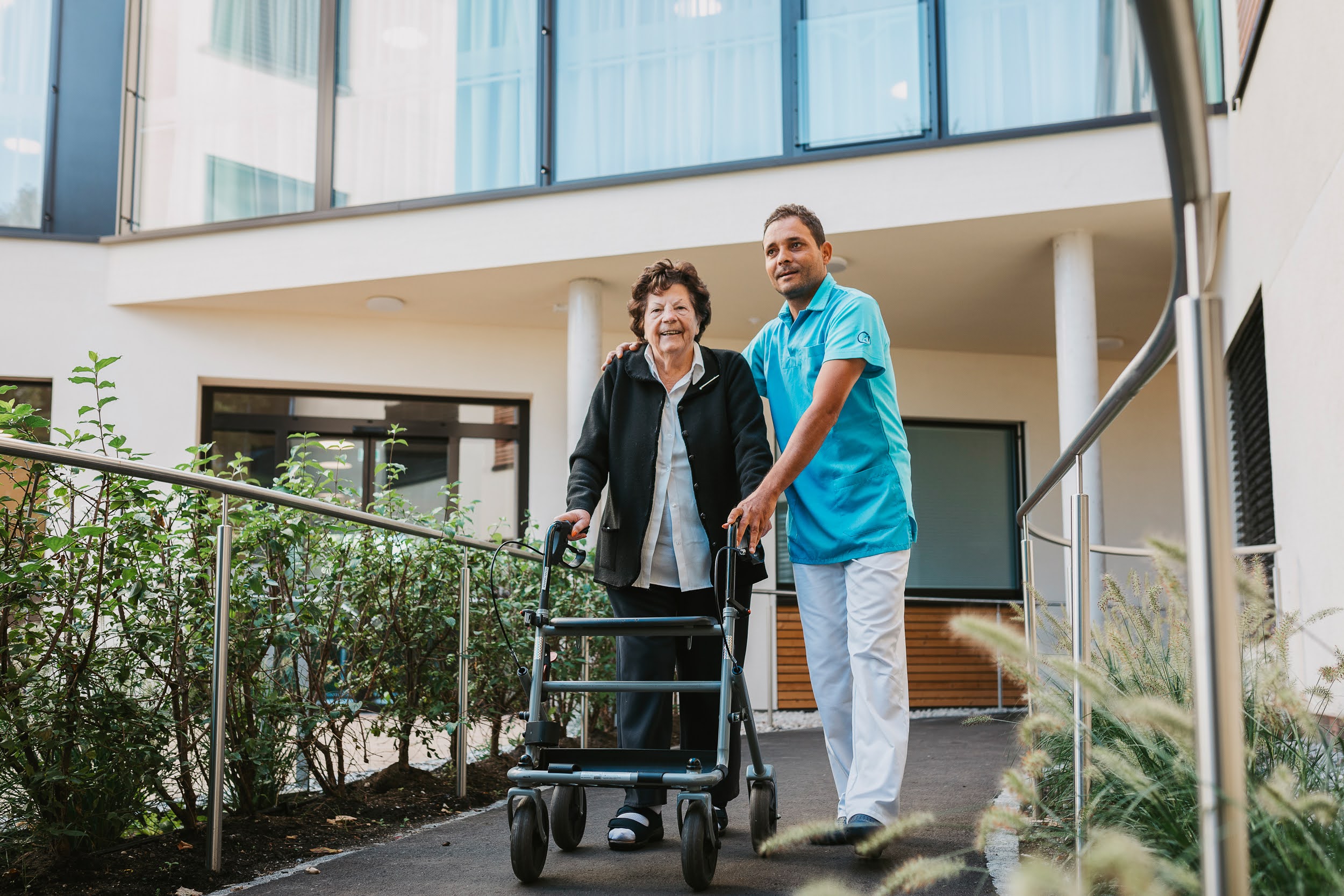 Auf dem Bild ist ein Pfleger des SeneCura Sozialzentrums Lauterach und eine ältere Frau mit einem Rollator zu sehen. 