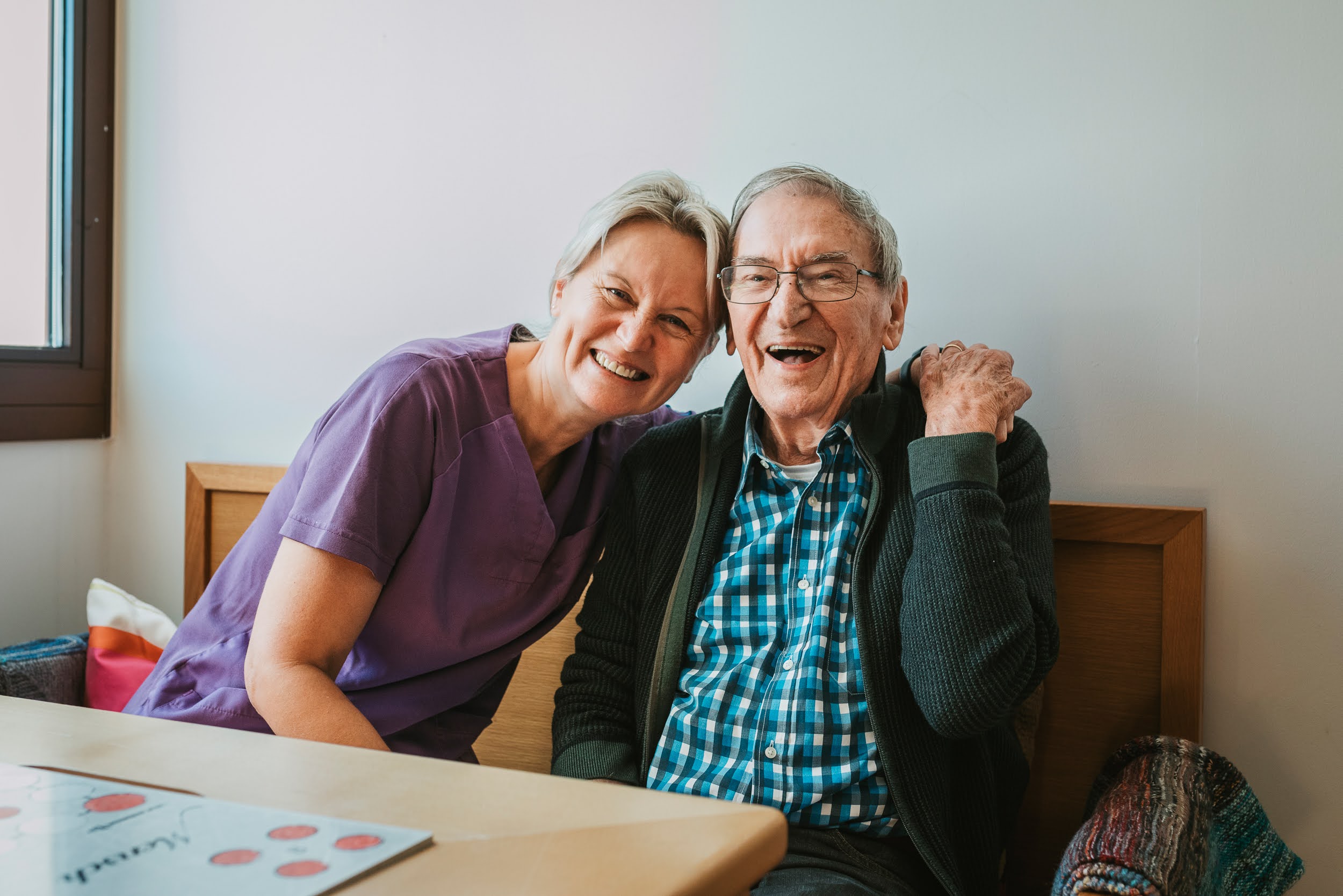 Auf dem Bild ist eine Pflegerin des SeneCura Sozialzentrums Lauterach zu sehen, die einen älteren Herren umarmt. Beide lachen.