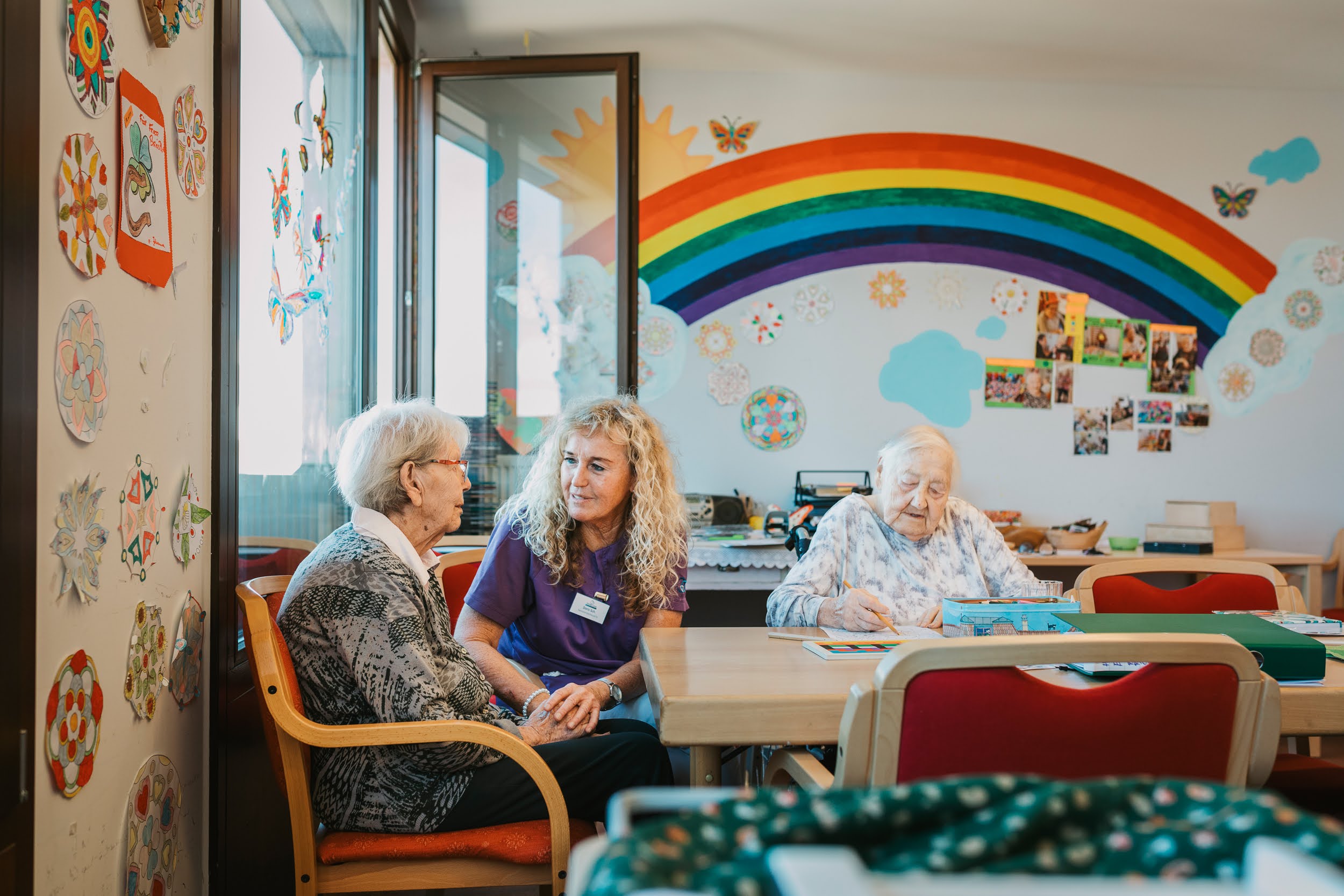 Auf dem Bild ist eine Pflegerin des SeneCura Sozialzentrum Lauterachs zu sehen, die gemeinsam it zwei älteren Frauen an einem Tisch sitzt. Sie hält die Hand einer Frau. Die andere Frau malt.