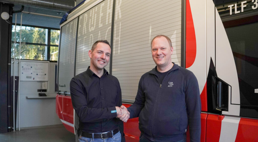 Das Bild zeigt zwei Männer, die vor einem Feuerwehrauto stehen und händeschüttelnd in die Kamera lächeln.