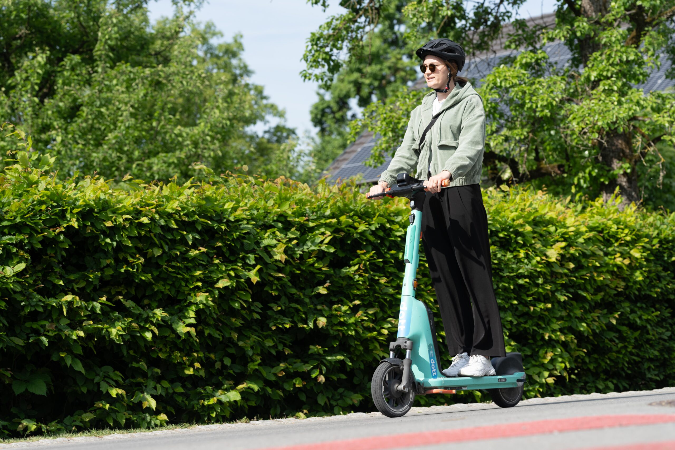Das Bild zeigt eine Frau, die mit Helm auf einer Straße mit einem Leih-E-Scooter der Marke DOTT fährt.