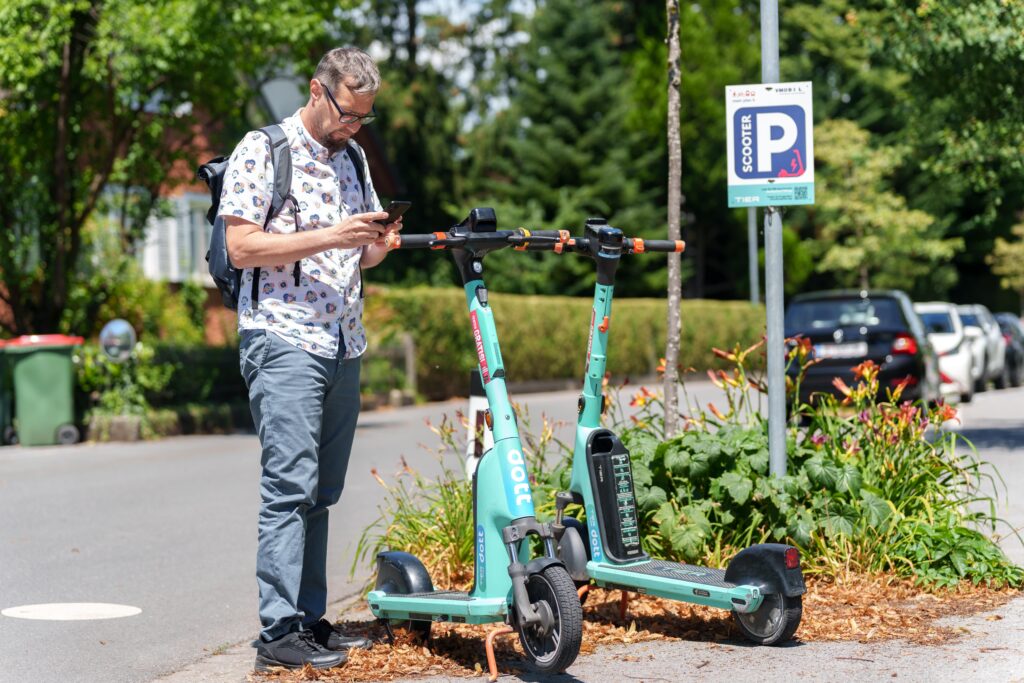 Das Bild zeigt einen Mann, der mit einem Smartphone in der Hand an einer Straße bei einem E-Scooter-Abstellplatz steht.