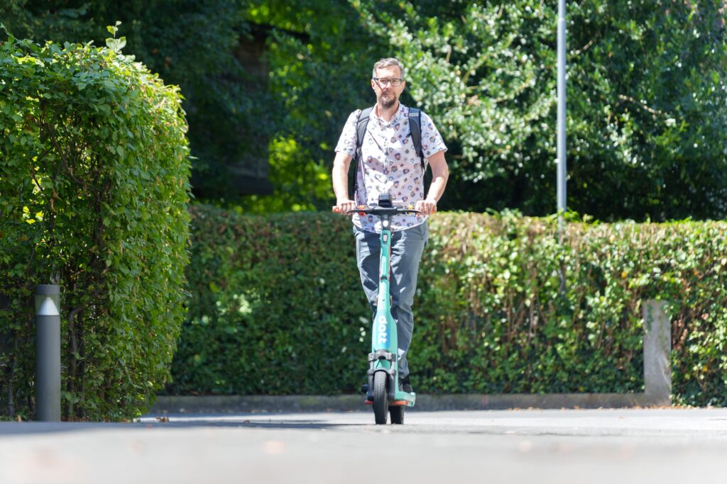 Das Bild zeigt einen Mann, der auf einer Straße mit einem Leih-E-Scooter der Marke DOTT fährt.