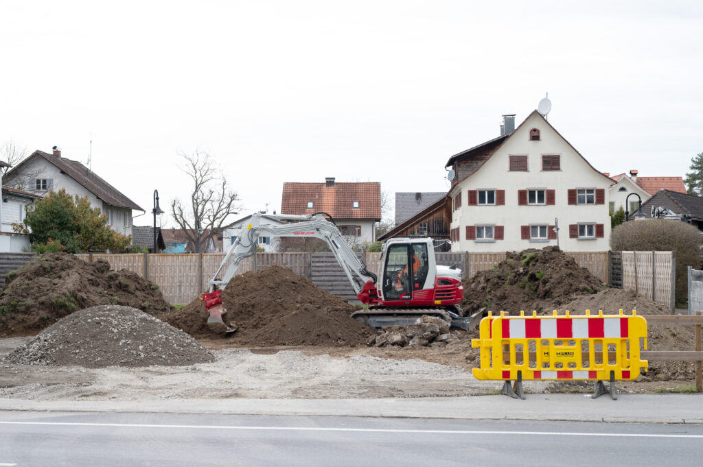 Das Bild zeigt eine Baustelle an der Bundesstraße in Lauterach mit Erdhaufen und einem Bagger.