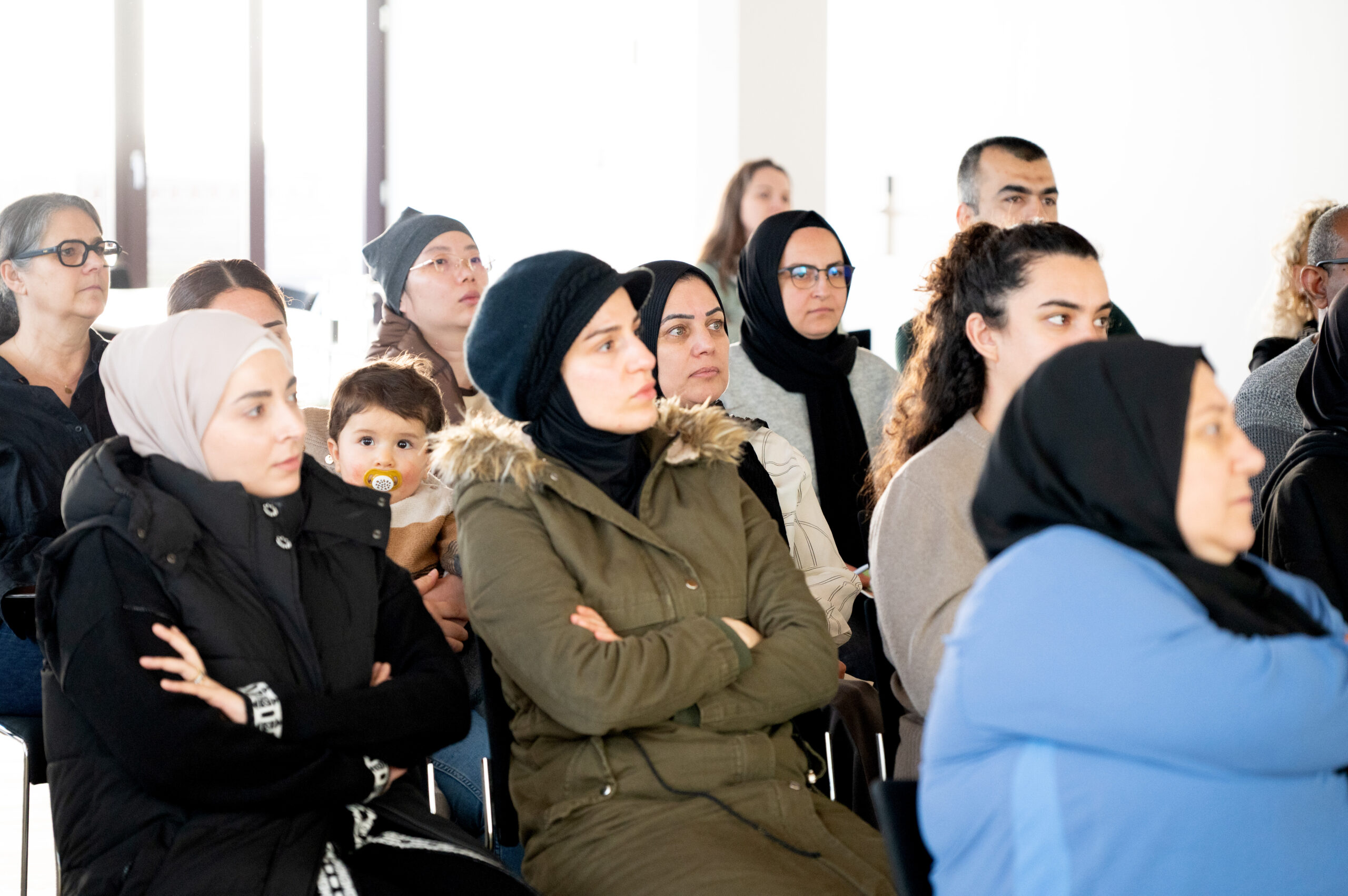Das Bild zeigt Frauen, ein Mann und ein Kleinkind, die im Publikum bei einer Veranstaltung sitzen.