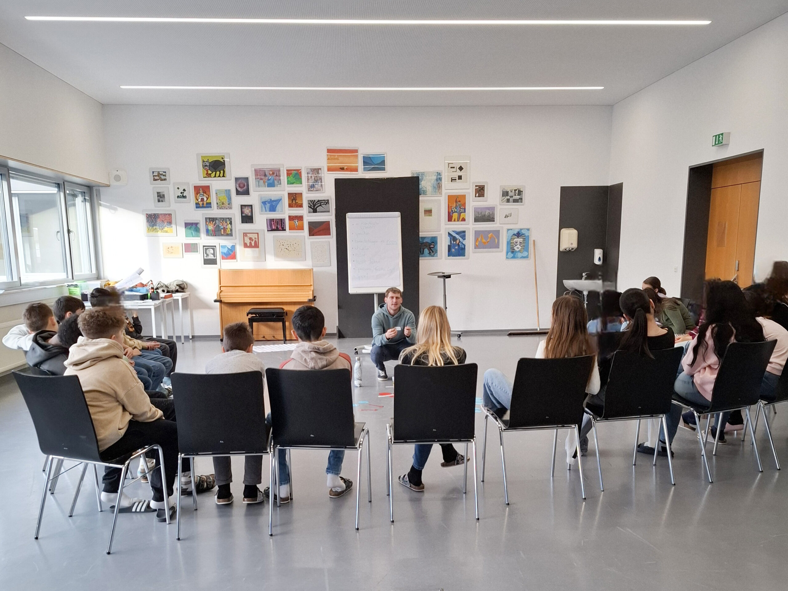 Das Bild zeigt eine Workshopsituation, in der eine Gruppe von Jugendlichen von hinten zu sehen ist, die in einer Reihe auf Stühlen in einem Klassenzimmer sitzt und einem Mann zuhört, der auf dem Boden kniet.