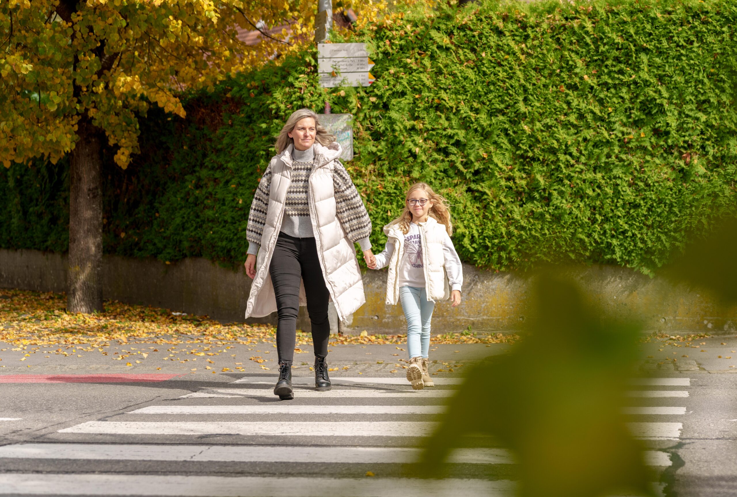 Das Bild zeigt eine Frau und ein Mädchen, die Hand in Hand über einen Zebrastreifen gehen.