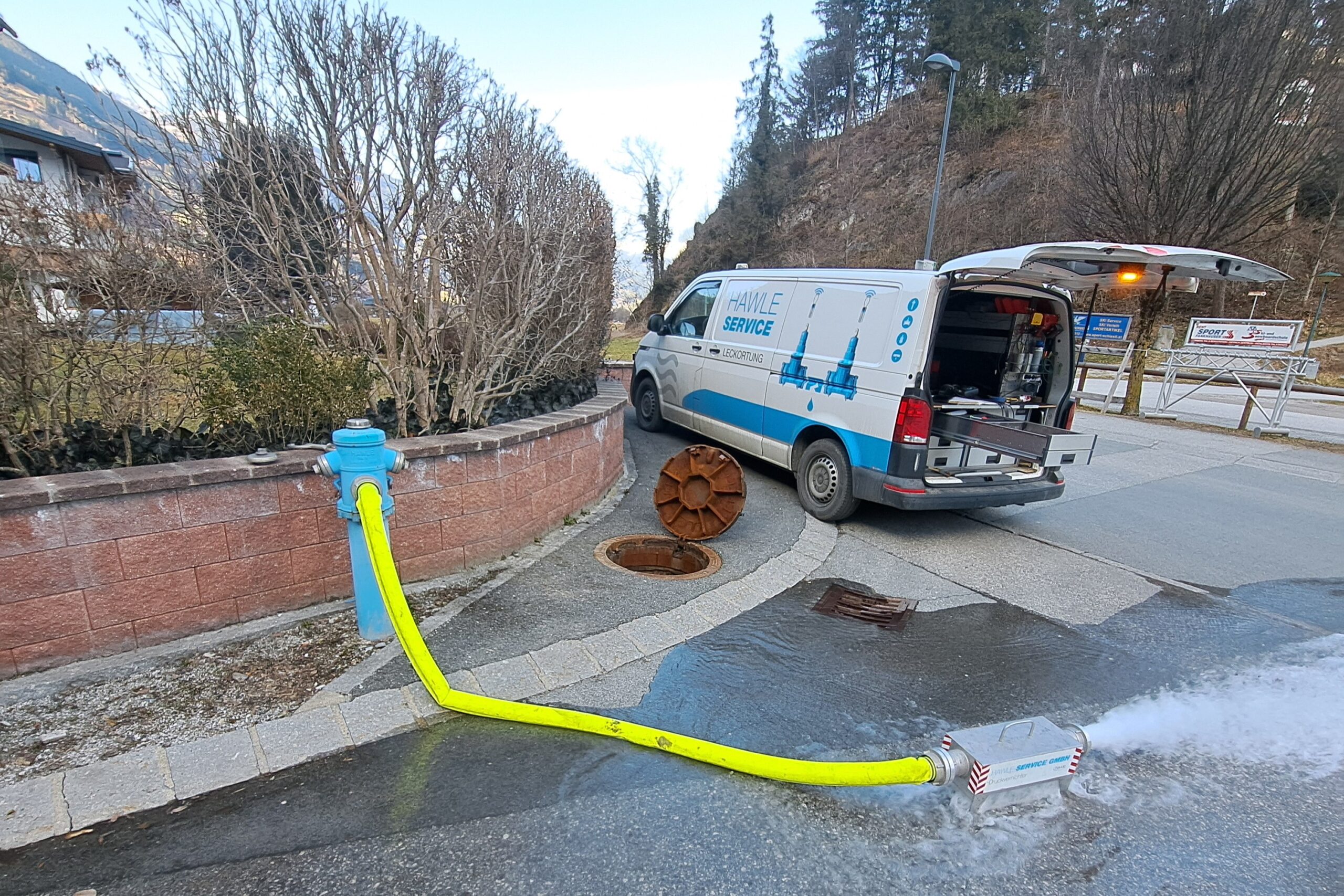Das Bild zeigt einen Transporter auf einer Straße und einen Hydrant, von dem über ein Schlauch Wasser auf die Straße abgeleitet wird.