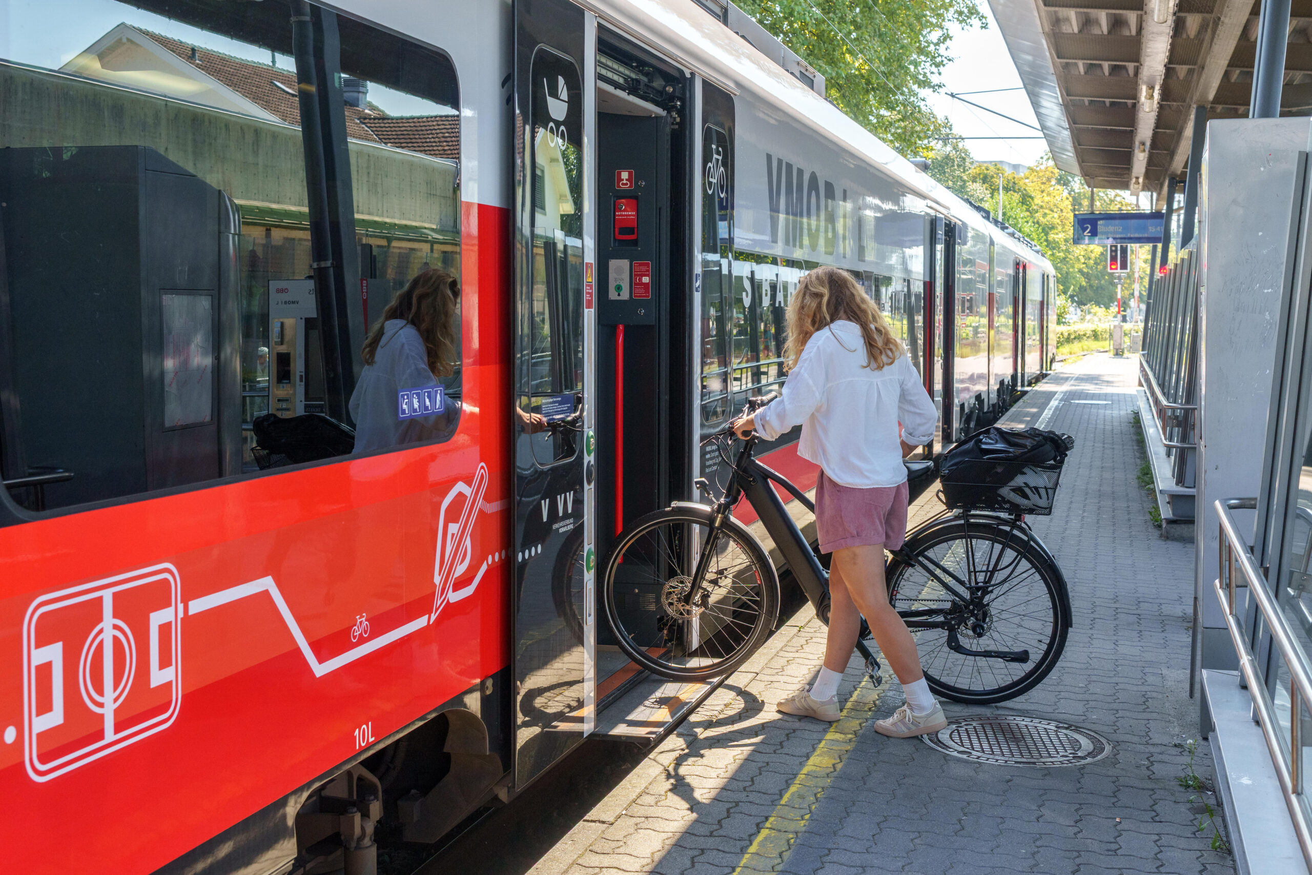 Das Bild zeigt eine Frau, die an einem Bahnhof mit einem Fahrrad in der Hand in einen Zug einsteigt.