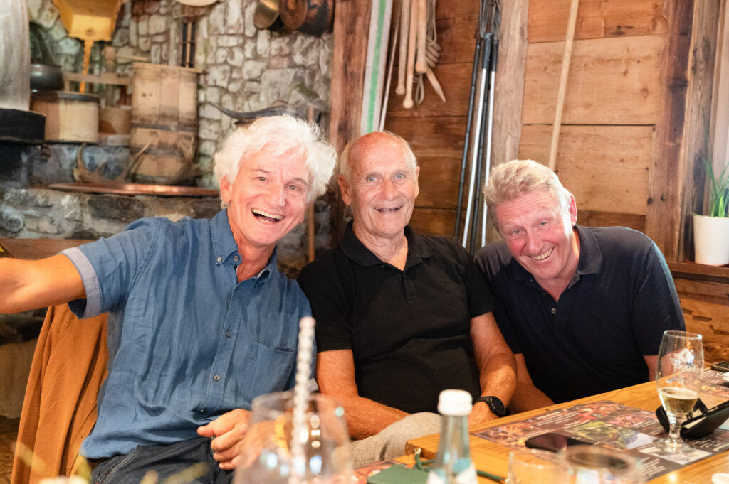 Drei Männer mit weißen Haaren sitzen nebeneinander an Holztischen und zeigen ein breites Lachen.