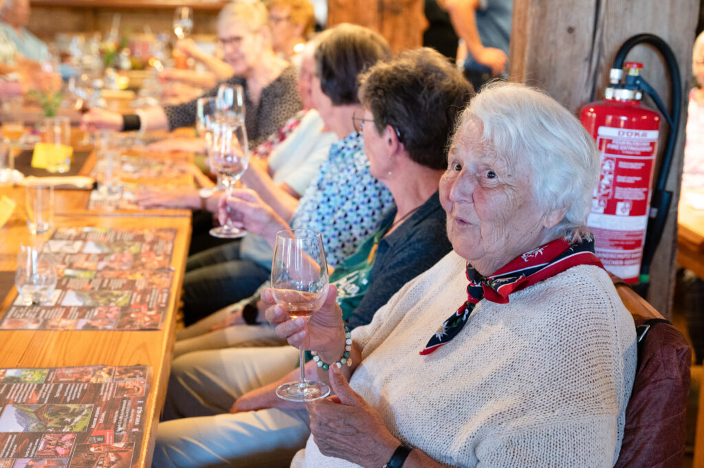 Mehrere ältere Personen sitzen nebeneinander an Holztischen. Eine Frau schaut in die Kamera und hält ein Weinglas mit Rosé in der Hand.