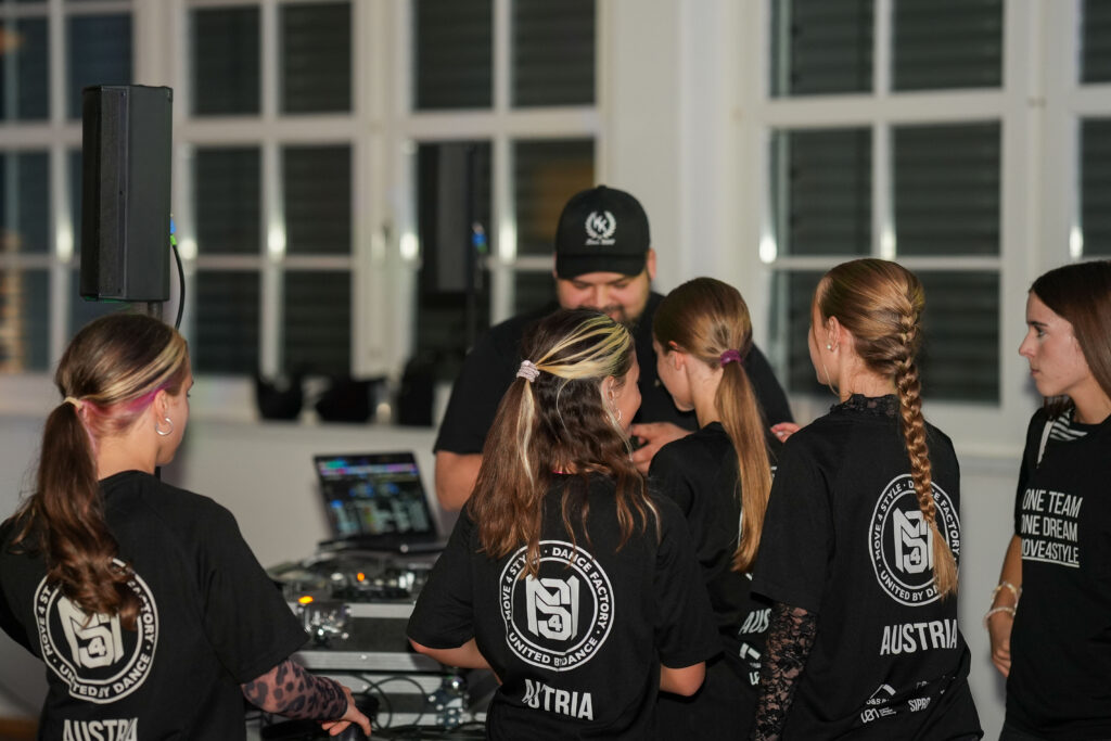 DJ Koloss steht am DJ-Pult und die Tanzgruppe wünscht sich einen Song von ihm.