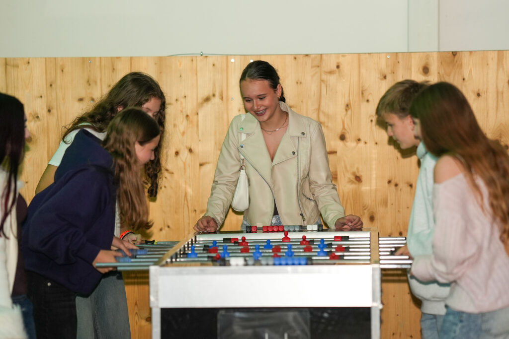 Die Jugend spielt Tischfußball in Teams zu zweit und ein Mädchen schaut zu.